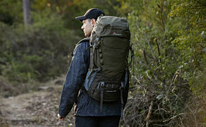 Une personne portant une veste et une casquette foncées est en randonnée avec une grande capacité de 55L, vert et noir, unisexe Sac à dos randonnée avec housse de pluie & H2O 1,65kg "MOUNTAINTOP - M5820II 55L" le long d'un sentier forestier. Une verdure dense entoure le sentier.