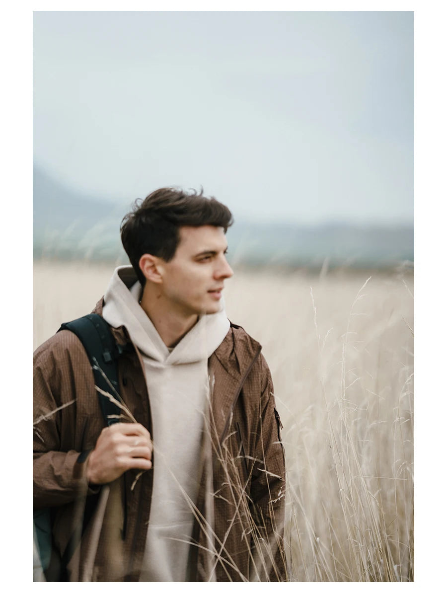 Un jeune homme portant une veste de randonnée coupe-vent et imperméable marron « SIMWOOD - SM130288 » et un sweat à capuche blanc se tient dans un champ d'herbes hautes, regardant sur le côté tout en tenant une sangle de sac à dos.