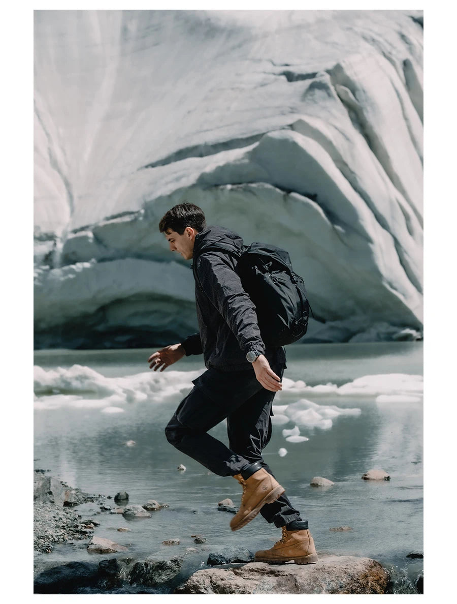 Une personne portant la veste de randonnée coupe-vent avec traitement hydrofuge pour homme Simwood « SIMWOOD - SM130288 », un pantalon noir et des bottes marron marche sur des rochers près d'un lac glaciaire avec des formations de glace en arrière-plan.