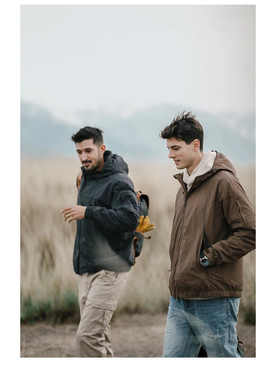 Deux hommes portant la veste de randonnée coupe-vent avec traitement hydrofuge pour homme Simwood « SIMWOOD - SM130288 » marchent dans un champ herbeux avec des montagnes en arrière-plan. Un homme porte un sac à dos avec des gants attachés.