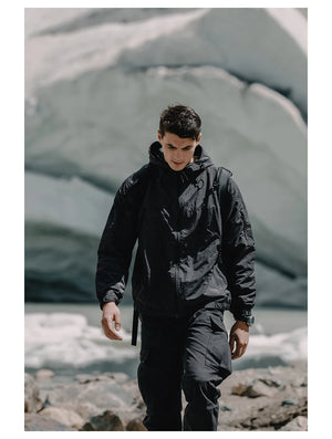 Une personne portant une veste de randonnée coupe-vent à capuche et un pantalon noirs Simwood - SM130288 marche à l'extérieur avec en toile de fond de gros rochers et d'un glacier.