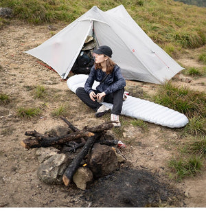 Une personne est assise sur un matelas de couchage près de la tente bivouac ultra-légère « 3F UL GEAR - Lanshan 2 pro », dotée d'une conception à simple paroi pesant 1,2 kg, profitant de la bonne ventilation fournie par l'installation en plein air sur le terrain herbeux à côté d'un feu de camp.